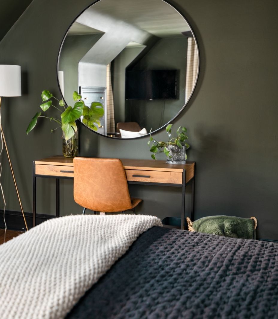 Schlafzimmer mit dunkelgrüner Wand, rundem Spiegel und einem Schreibtisch mit Pflanzen
